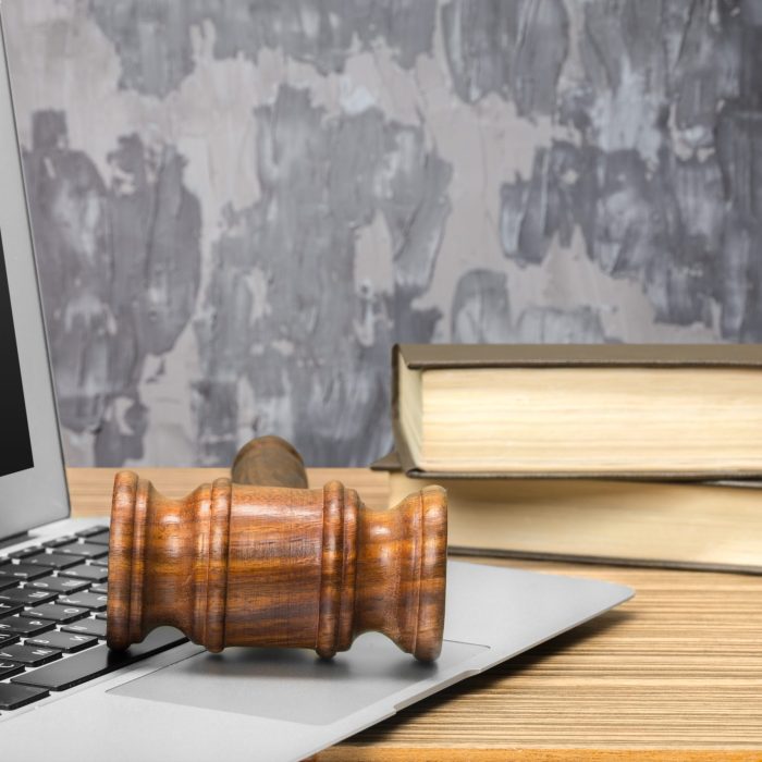 Drewniany młotek sędziowski leżący na klawiaturze laptopa, obok stos książek na drewnianym stole.